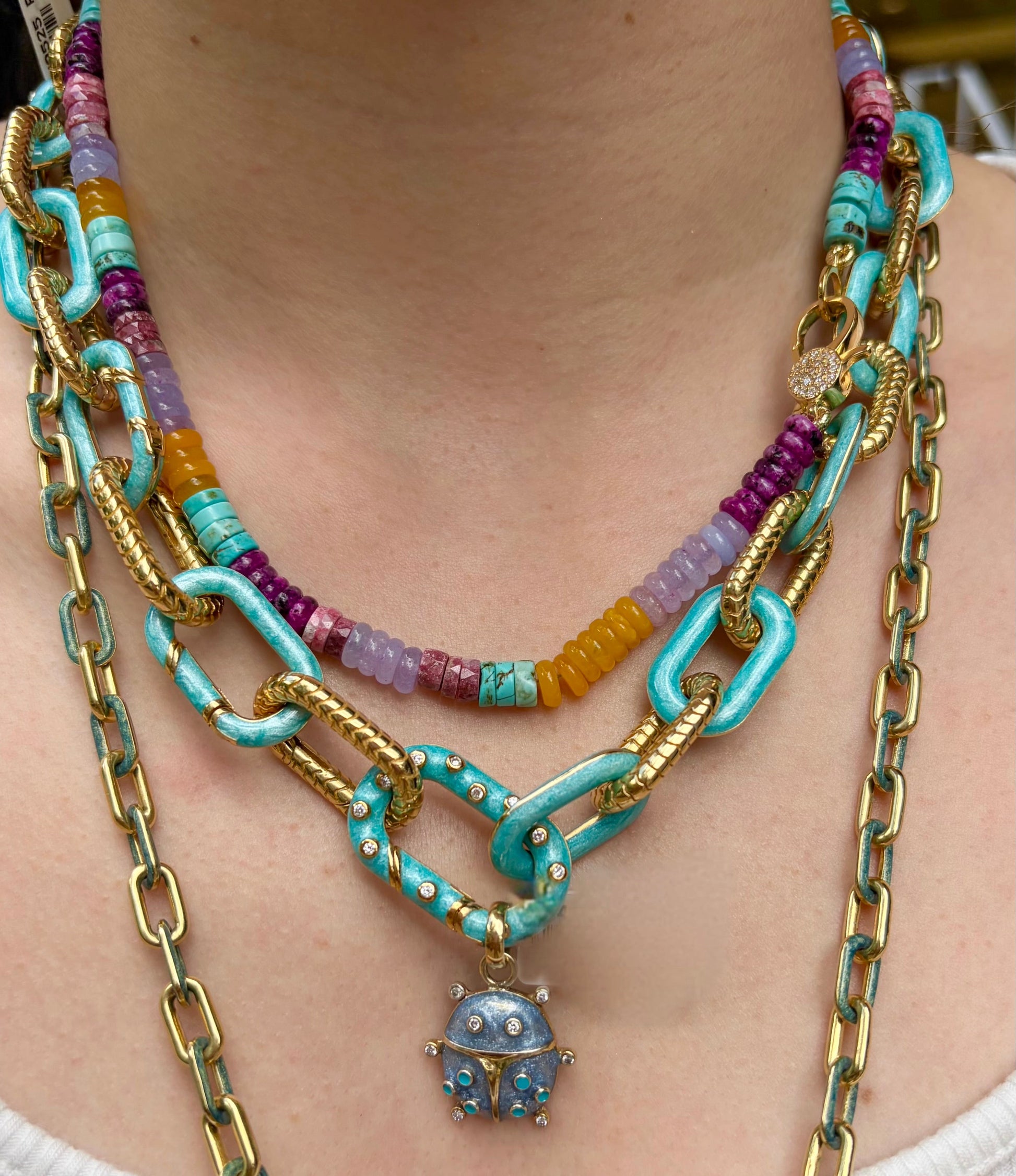 Colorful beaded and chain necklaces with a blue pendant on a person wearing a white top.